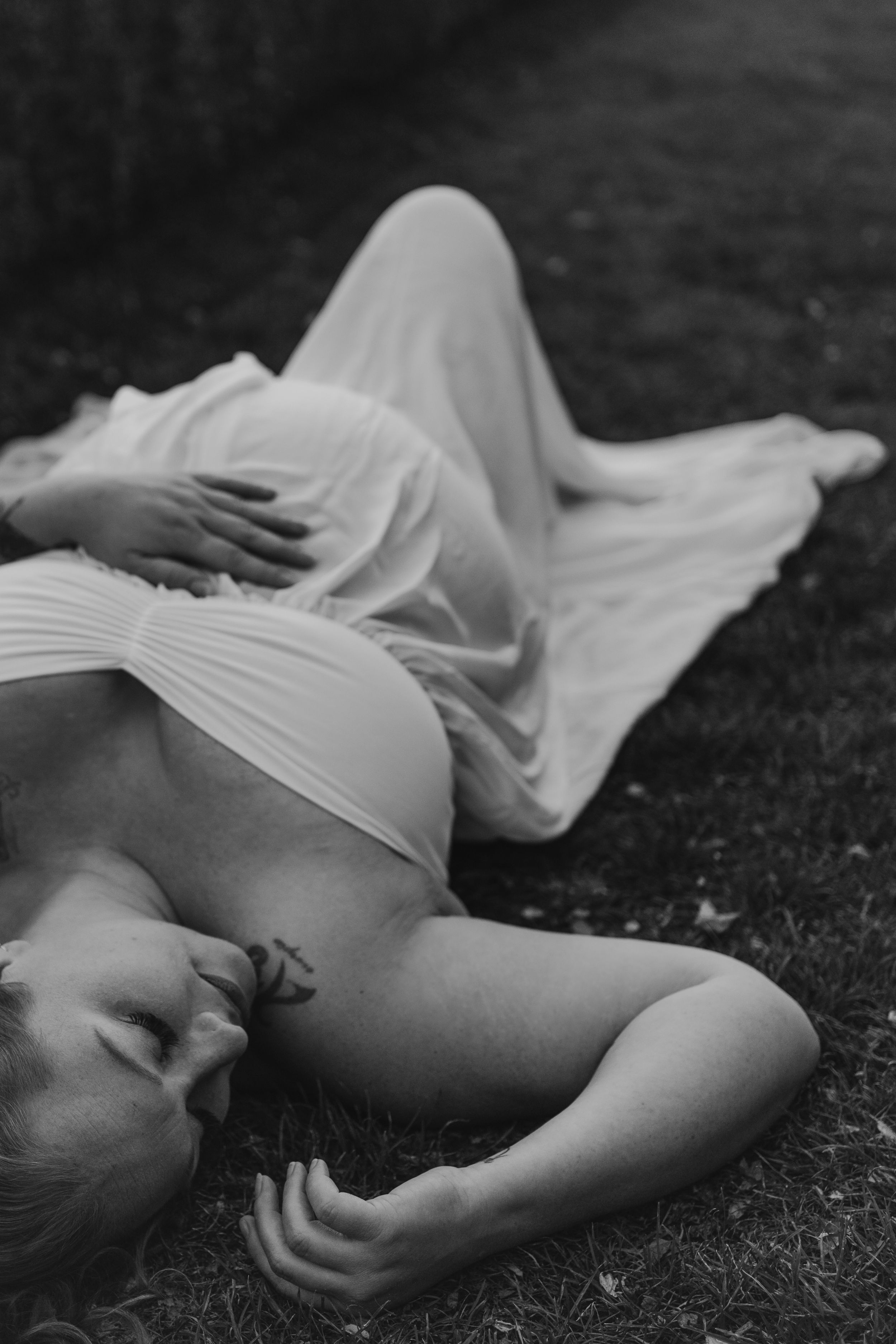 Artistic black and white photo of someone reclining on grass in flowing white fabric.