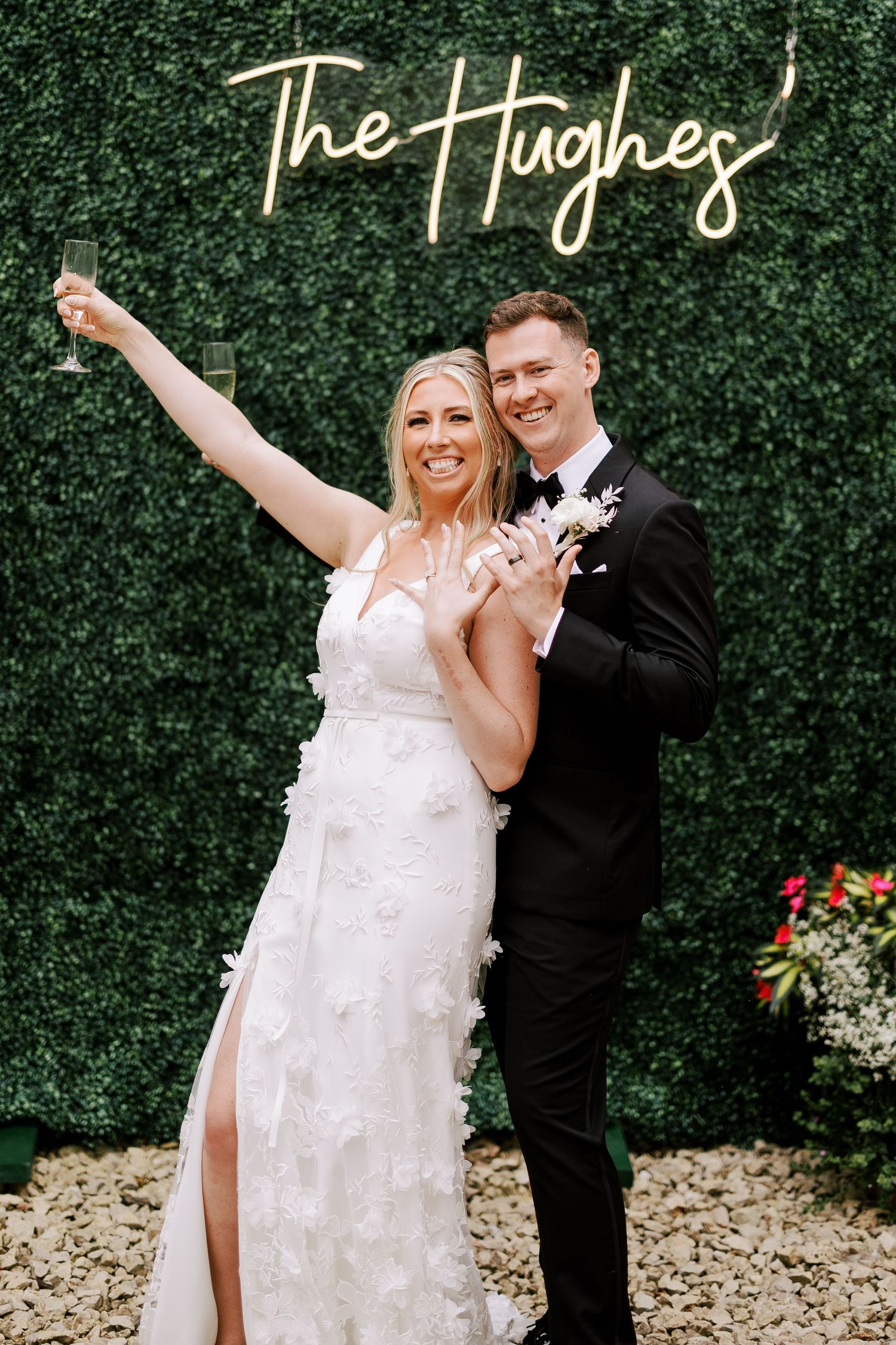 Wedding couple poses in front of green hedge wall with illuminated name sign.