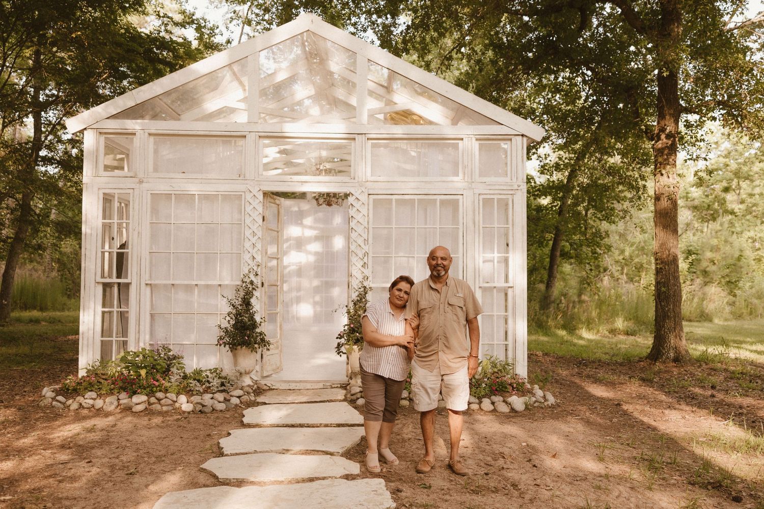 White greenhouse-style garden structure with decorative details set in a wooded outdoor setting.