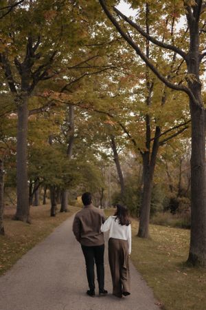 Two people walk hand in hand down a tree-lined path with golden fall foliage.