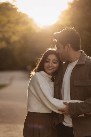Romantic couple embracing in warm sunset light with woman in white sweater and man in brown jacket.