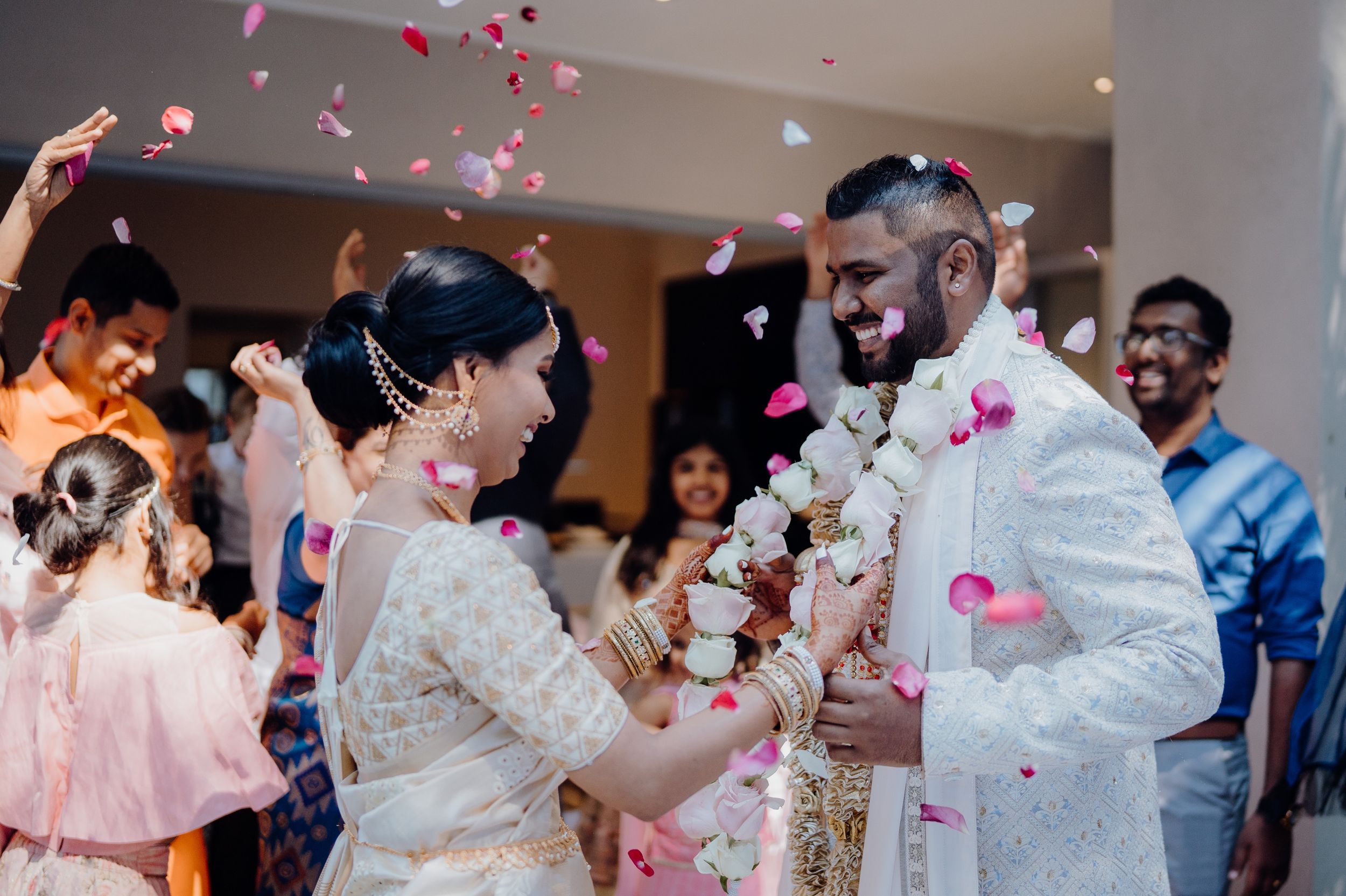 Pink flower petals fall during a traditional wedding ceremony as couple exchanges garlands in an indoor celebration.
