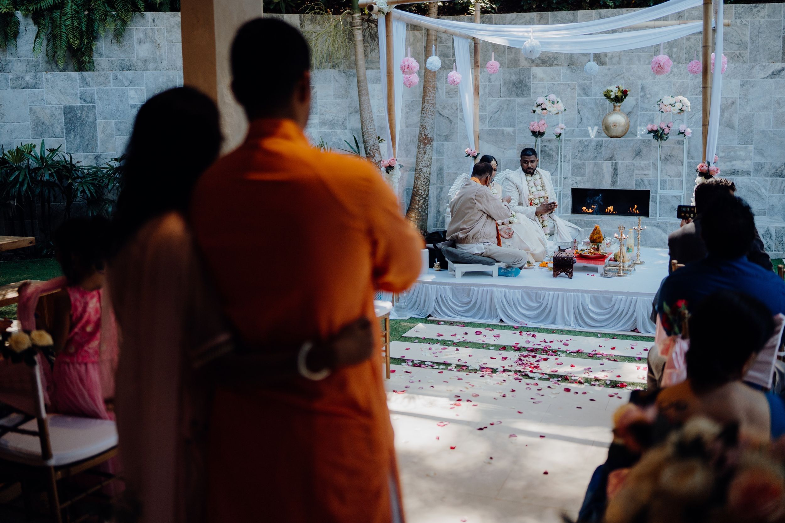 Guests observe a traditional outdoor wedding ceremony under white canopy with rose petals scattered around.