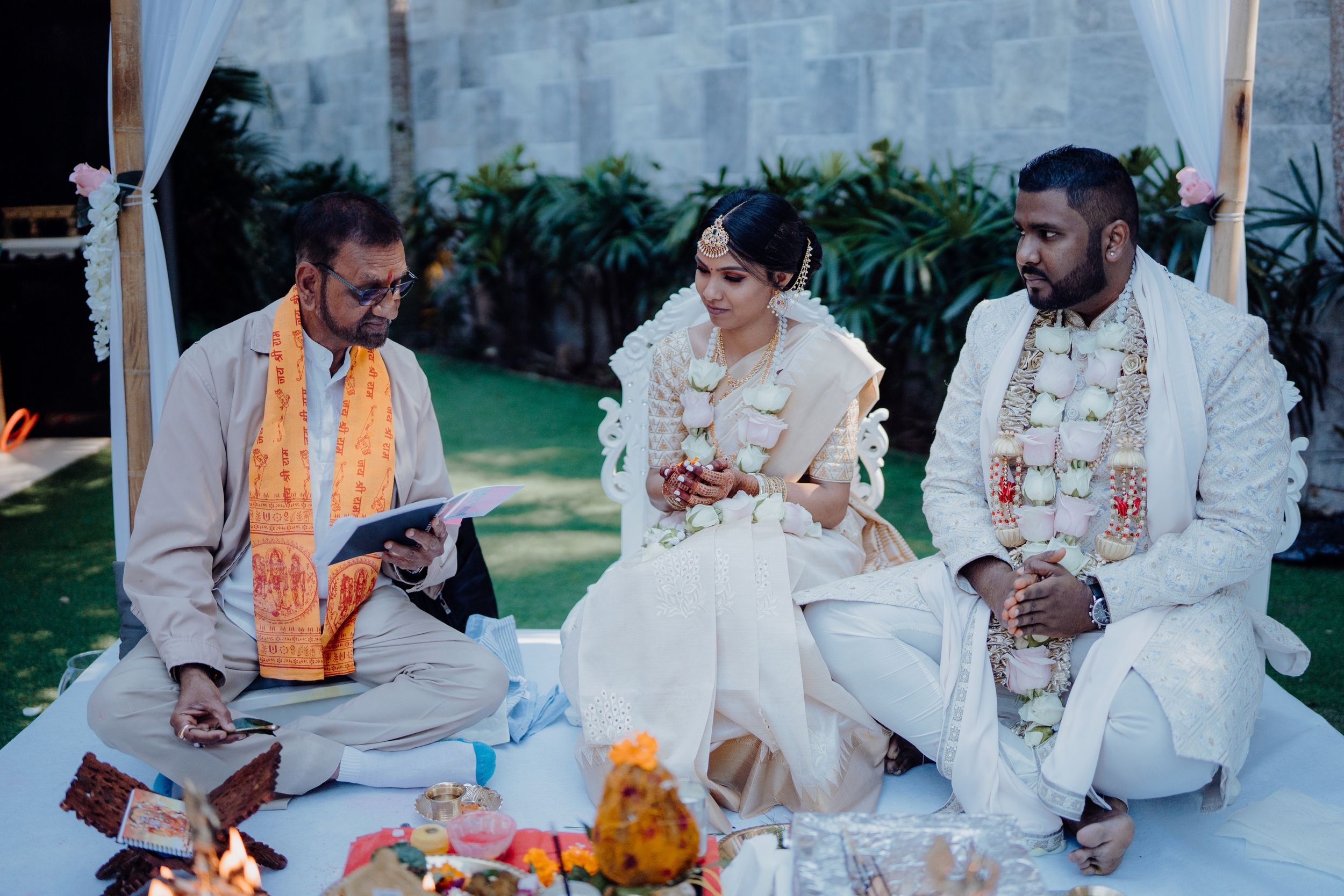 Traditional wedding ceremony participants seated on floor with ritual items and orange decorative elements.