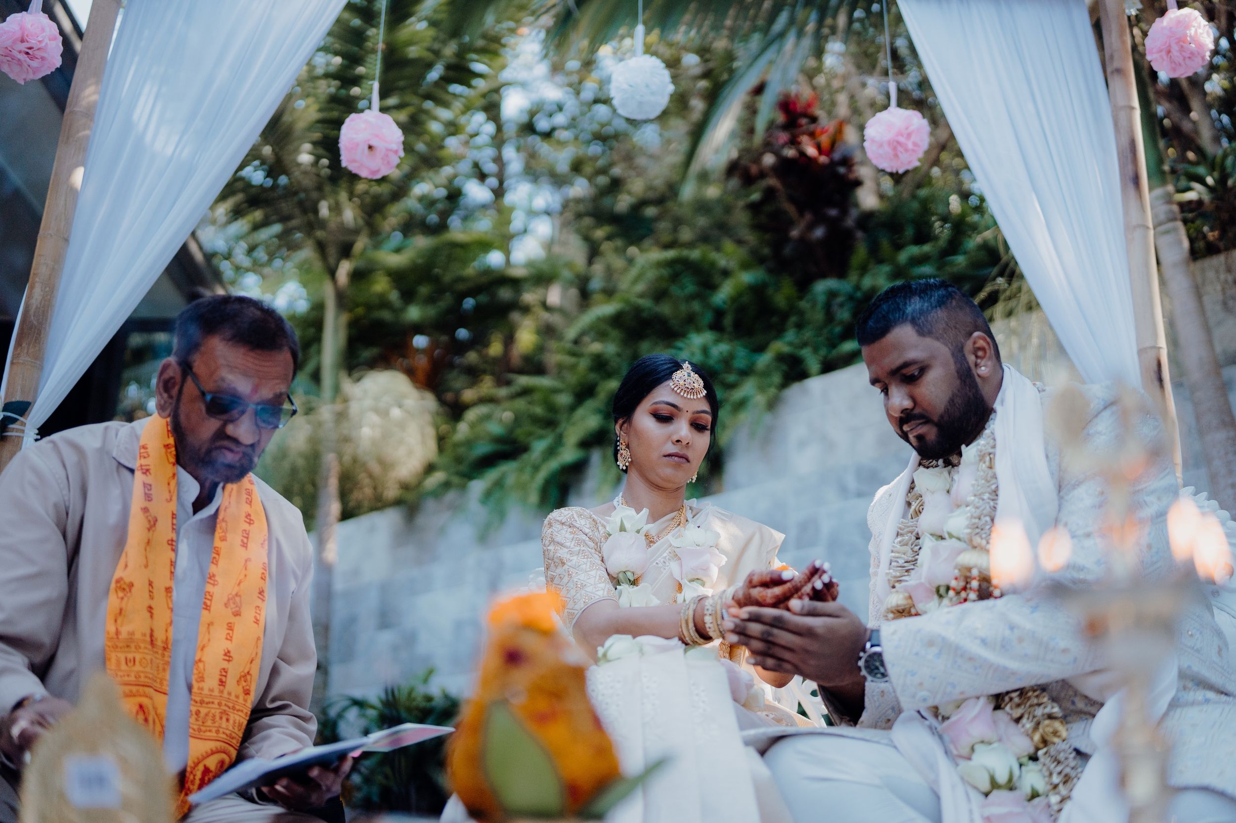 Wedding ceremony under white draped canopy with pink lanterns and tropical foliage.
