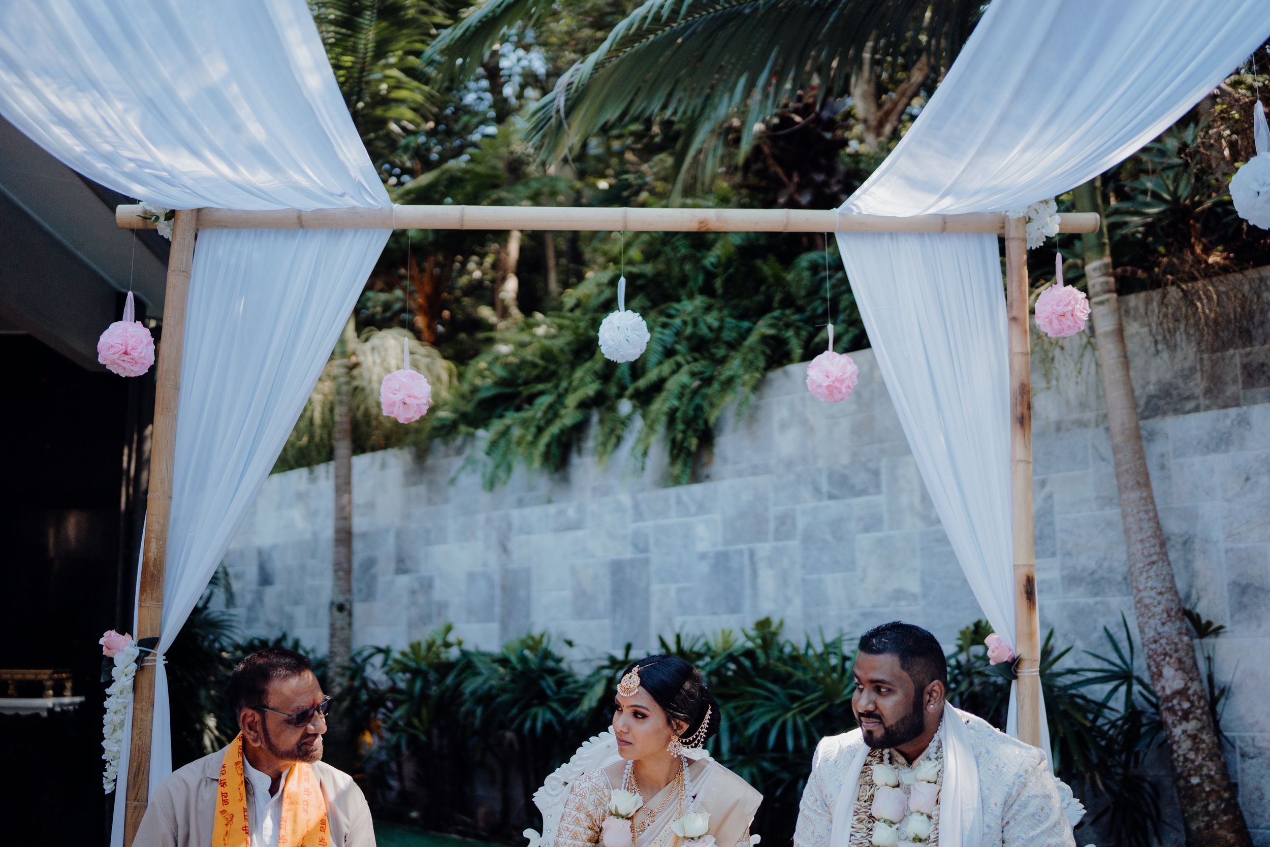 Outdoor wedding setup with white drapes, pink lanterns, and lush greenery backdrop.