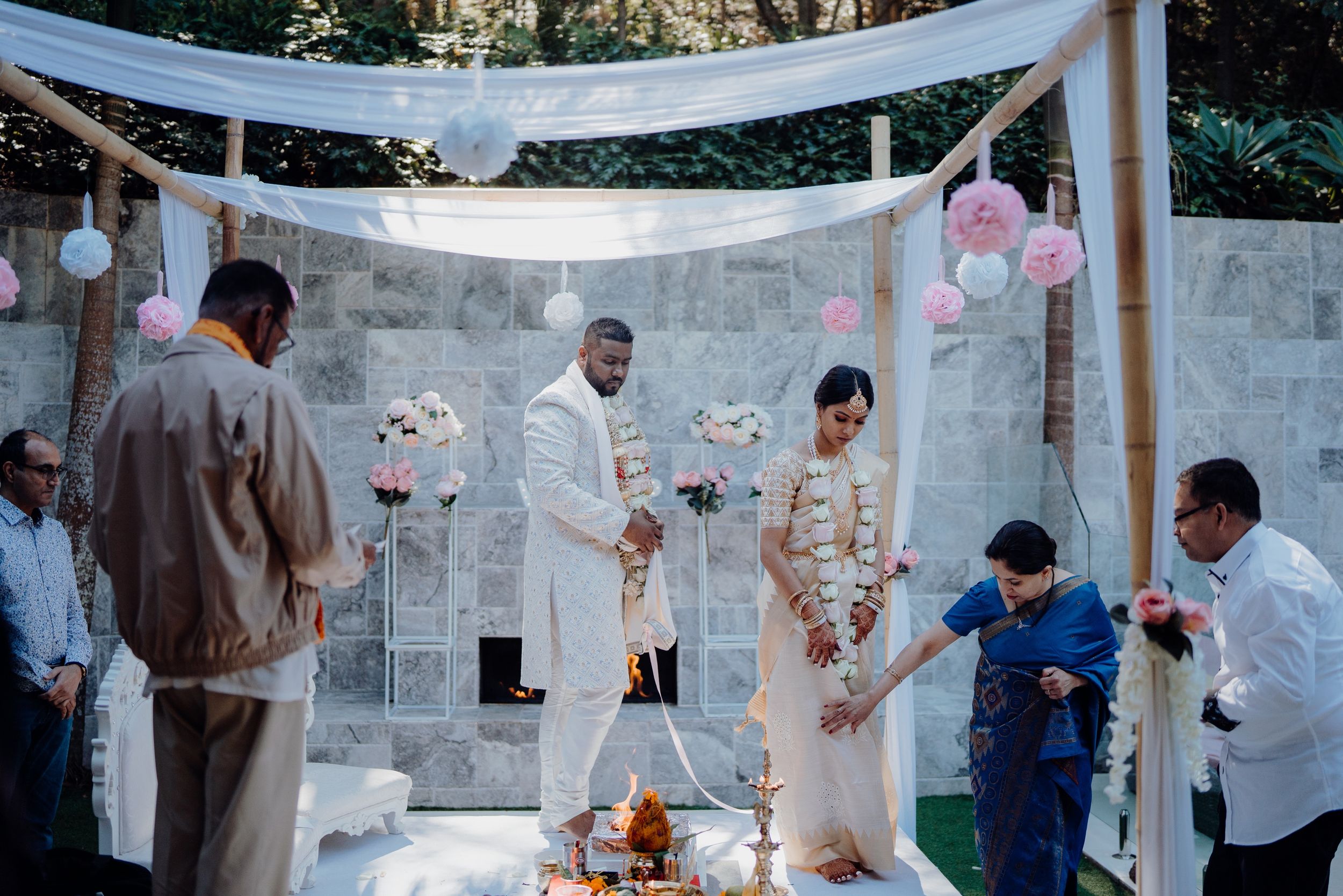 Outdoor wedding ceremony with white drapes and pink decorations against a stone wall.
