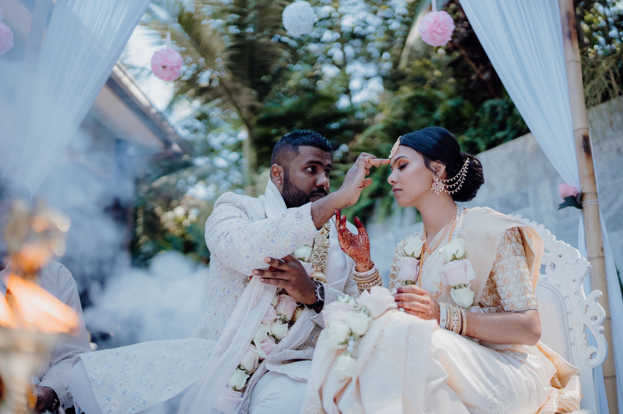 A couple participates in a traditional wedding ceremony under a draped canopy with hazy smoke.