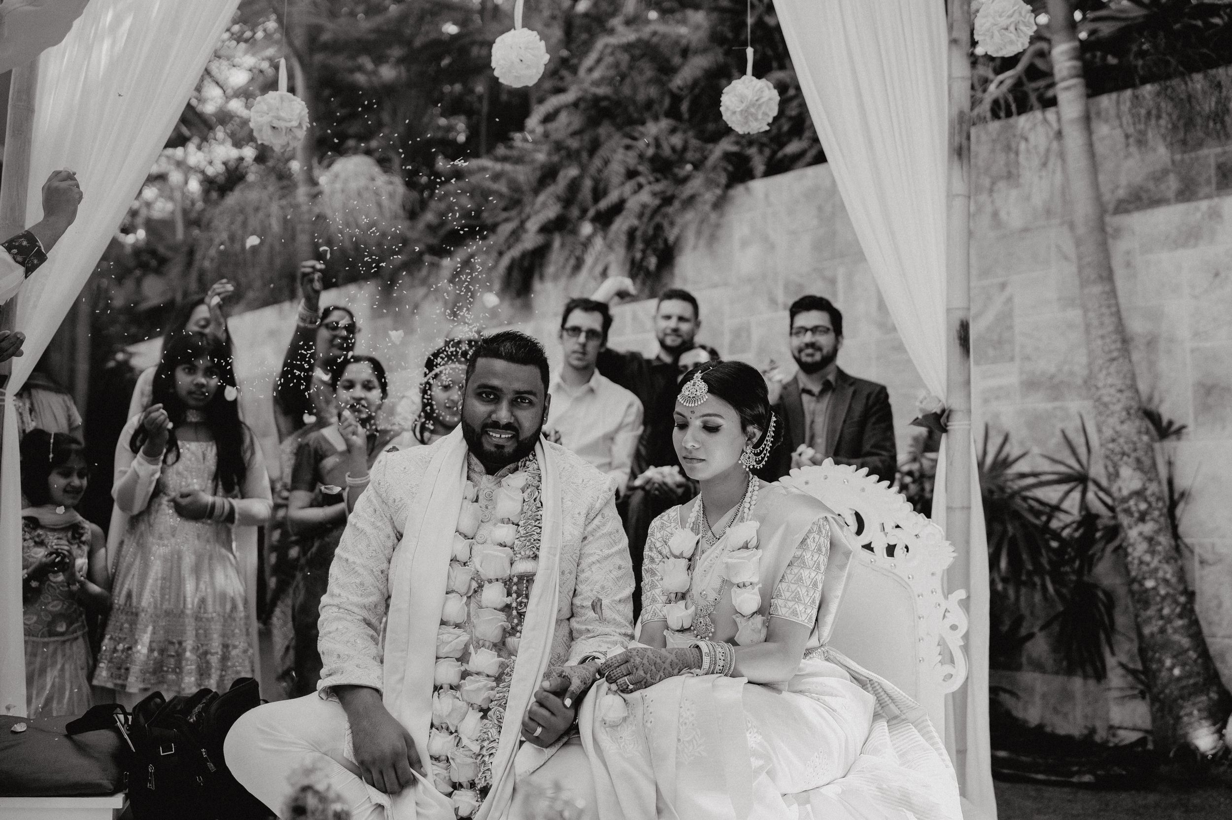Newlyweds sit together during a black and white wedding ceremony under white fabric draping and hanging lights.