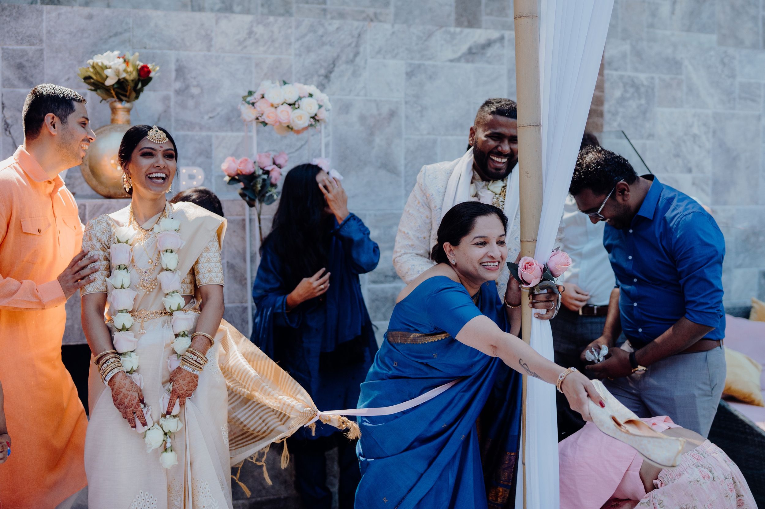 Wedding guests interact during an outdoor ceremony with white draping and stone wall backdrop.