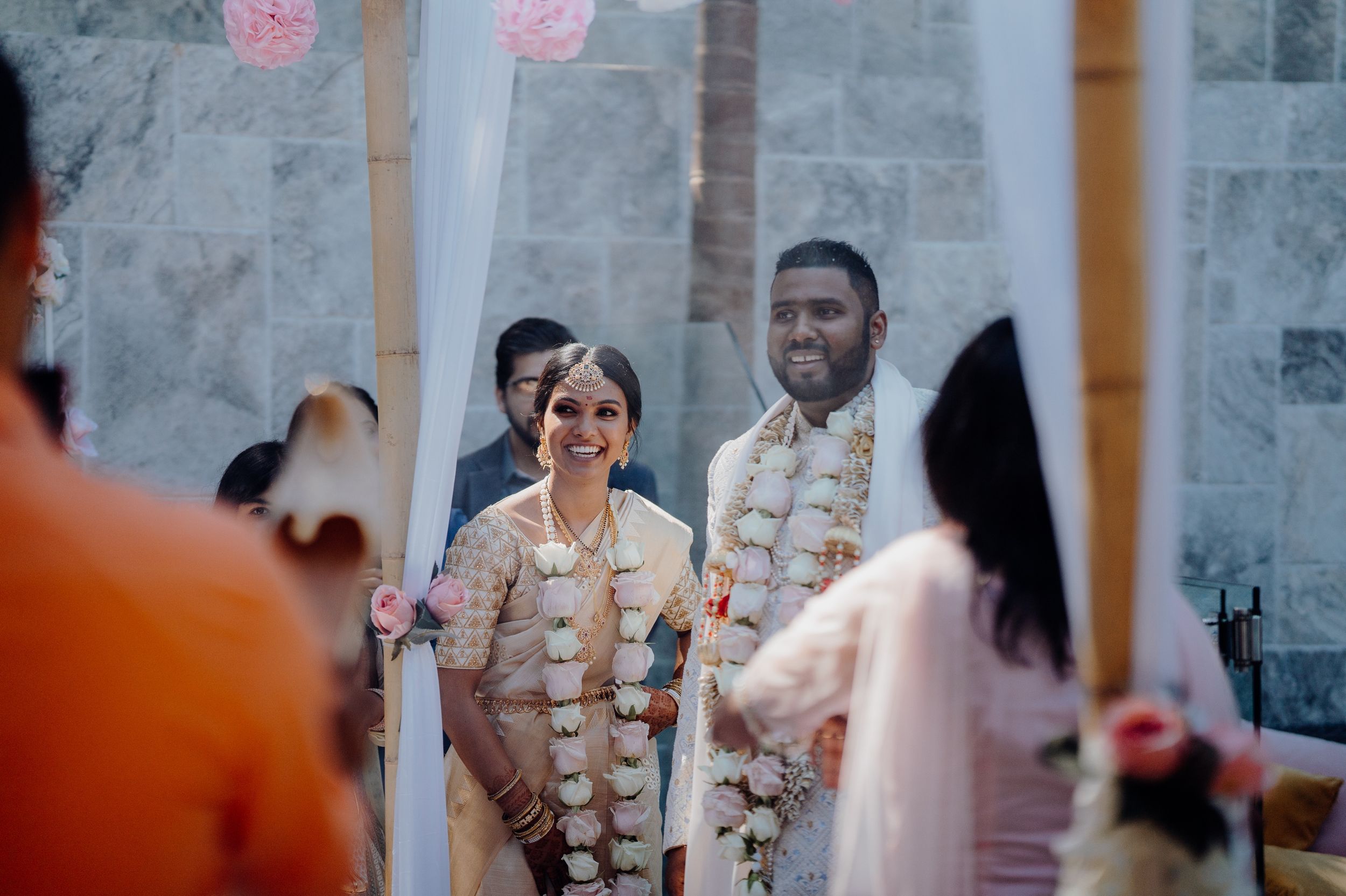 Couple wearing floral garlands celebrate during their wedding ceremony against a grey stone wall.