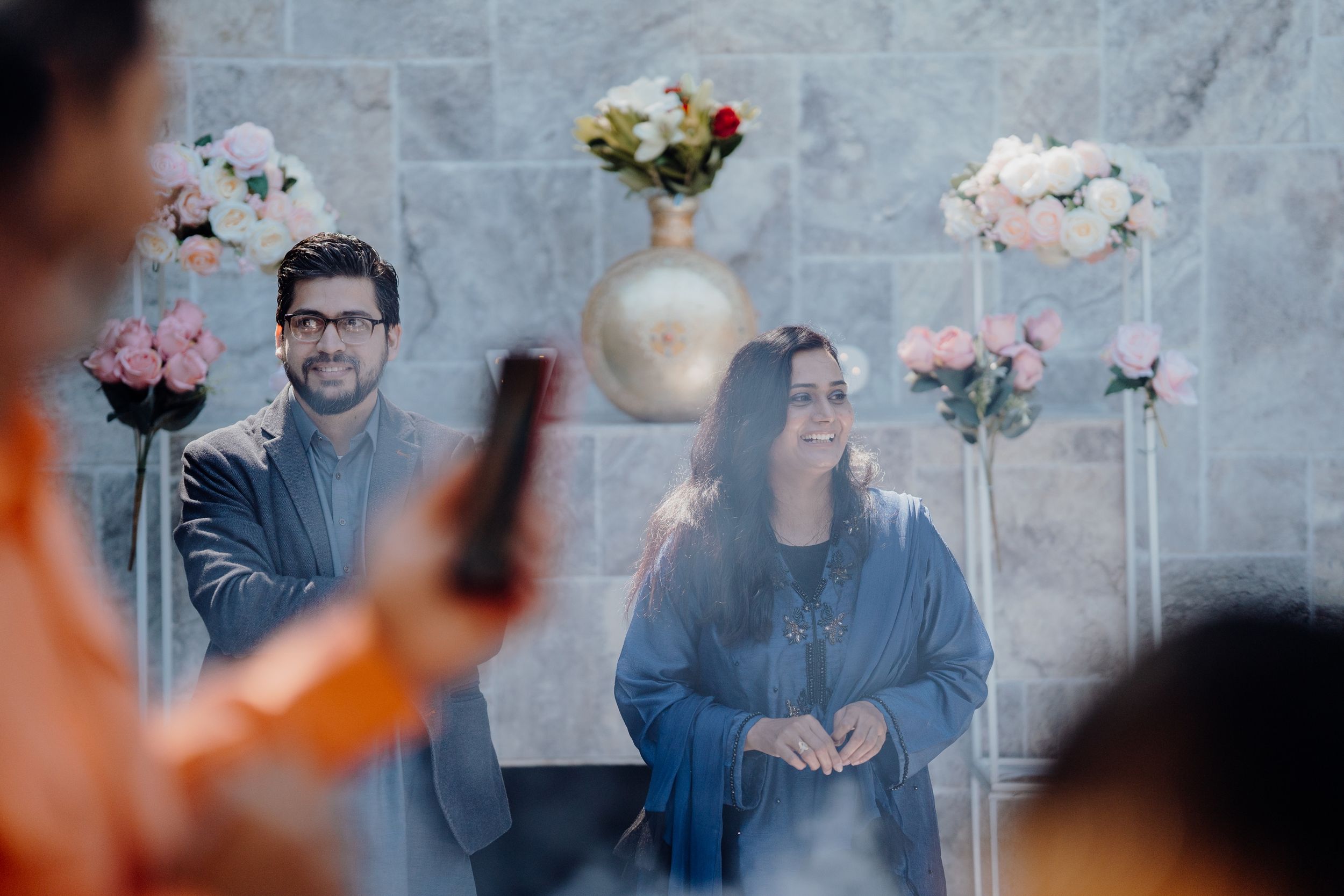 Wedding guests gather around floral decorations against a stone wall backdrop.