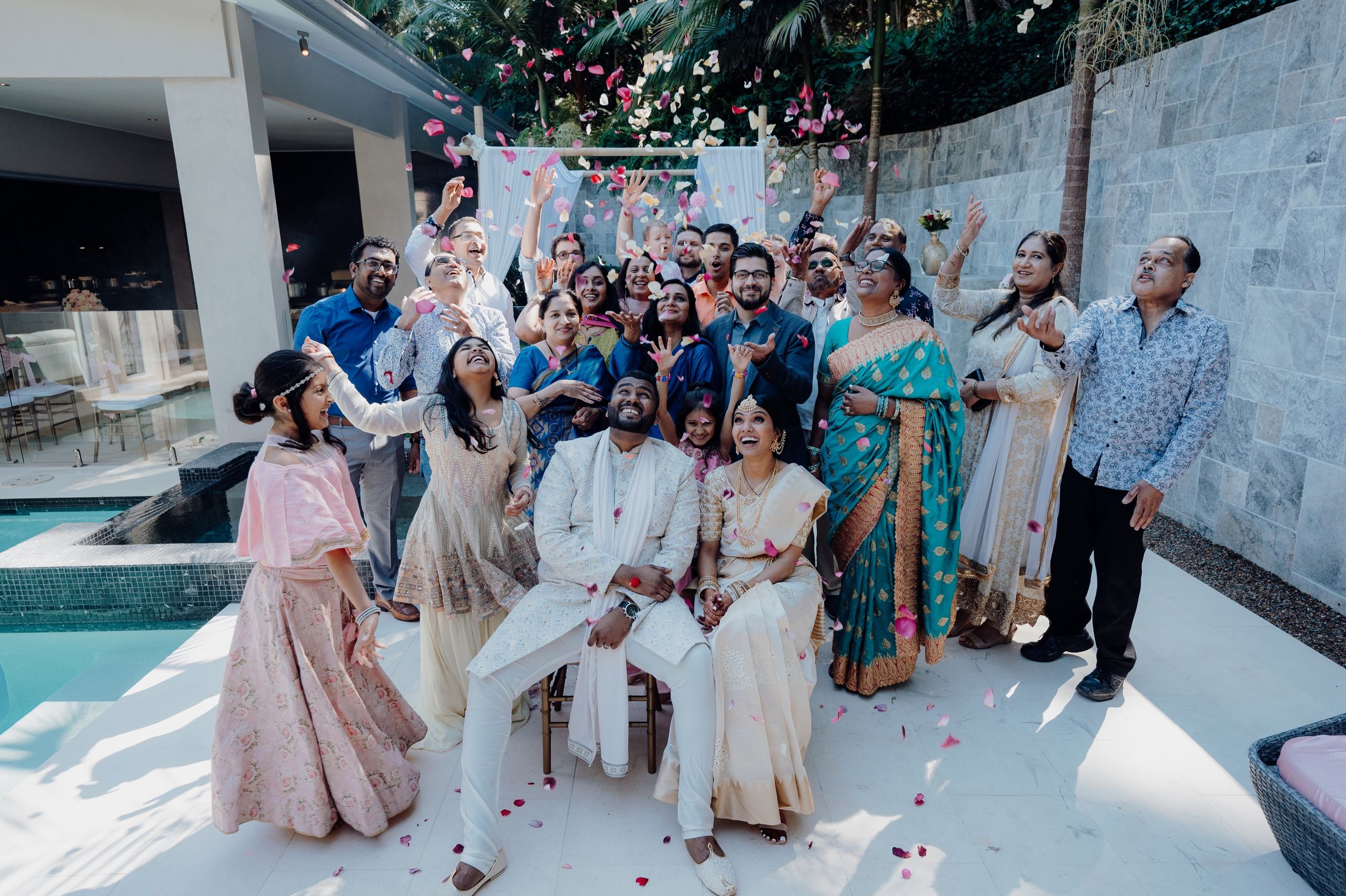 Wedding guests celebrate joyfully around the couple by a poolside setting.