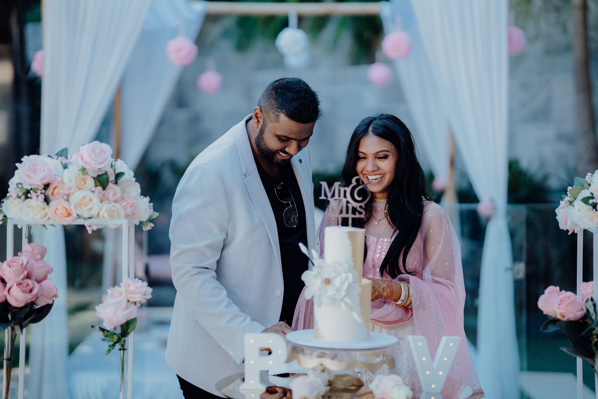 Couple cutting their elegant white wedding cake under a draped white canopy with floral decorations.