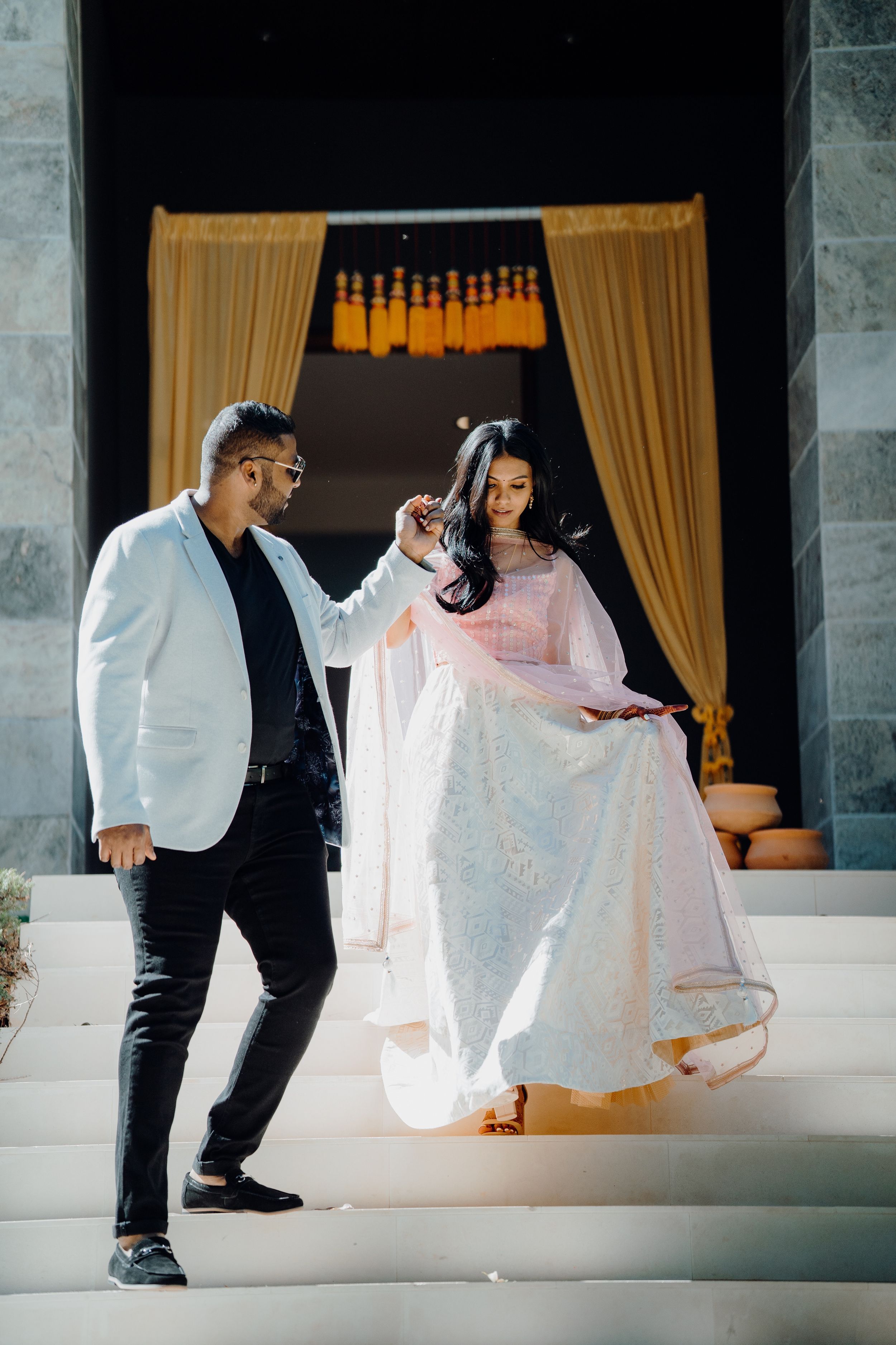 Couple descending white stairs in formal attire with golden curtains in background.