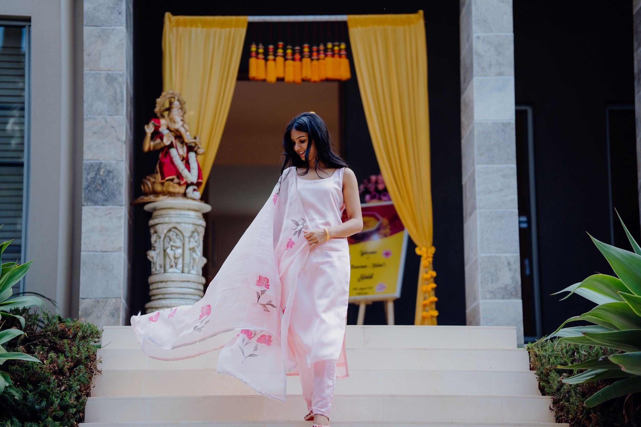 Pink floral dress against yellow curtains and traditional deity statue in entrance.