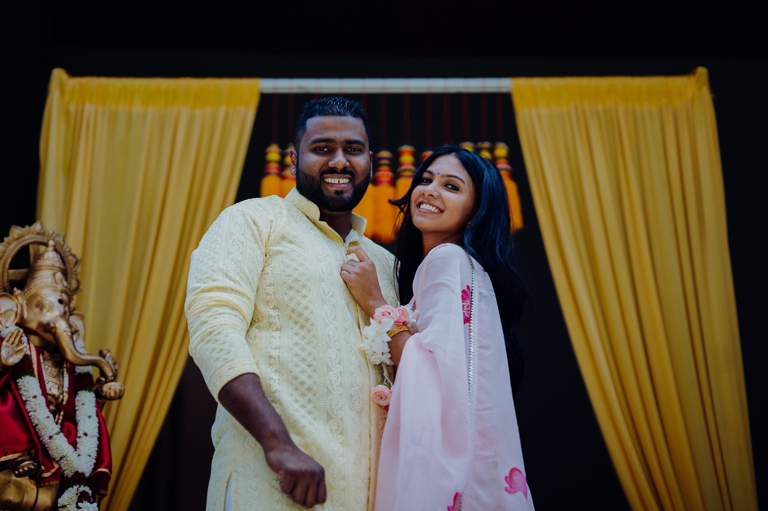 Couple in traditional Indian attire celebrating in front of yellow backdrop with decorative lighting.