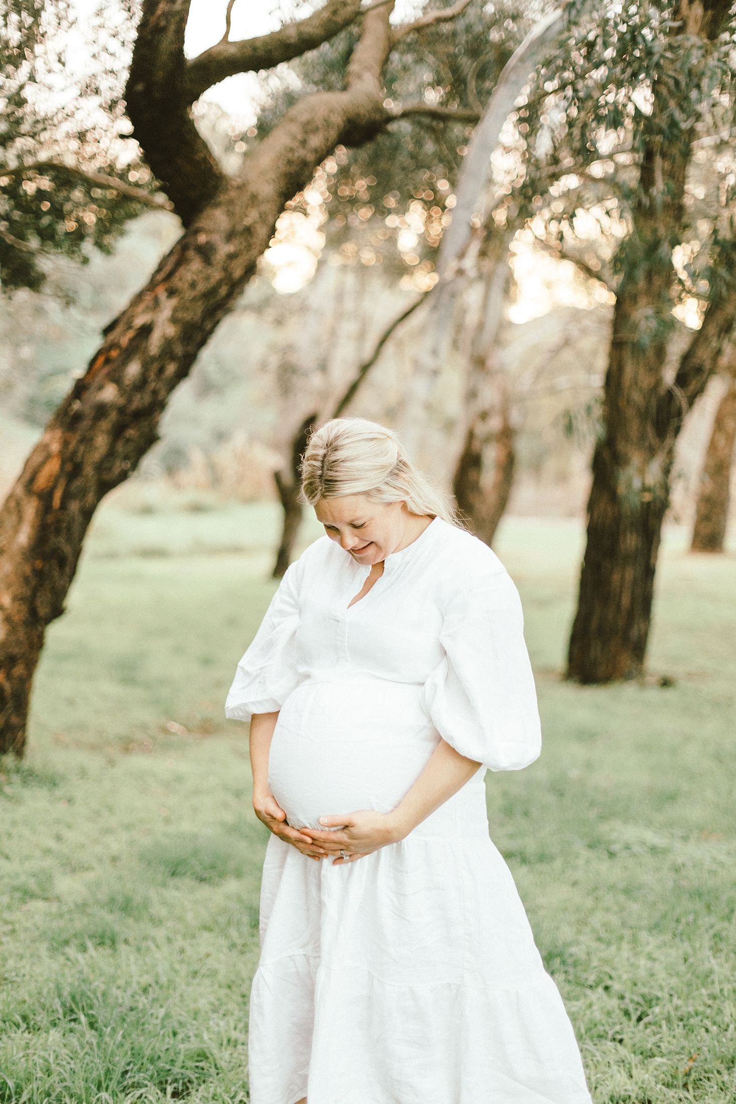 Maternity photos in a natural setting with trees showing woman in flowing white dress touching her baby bump.