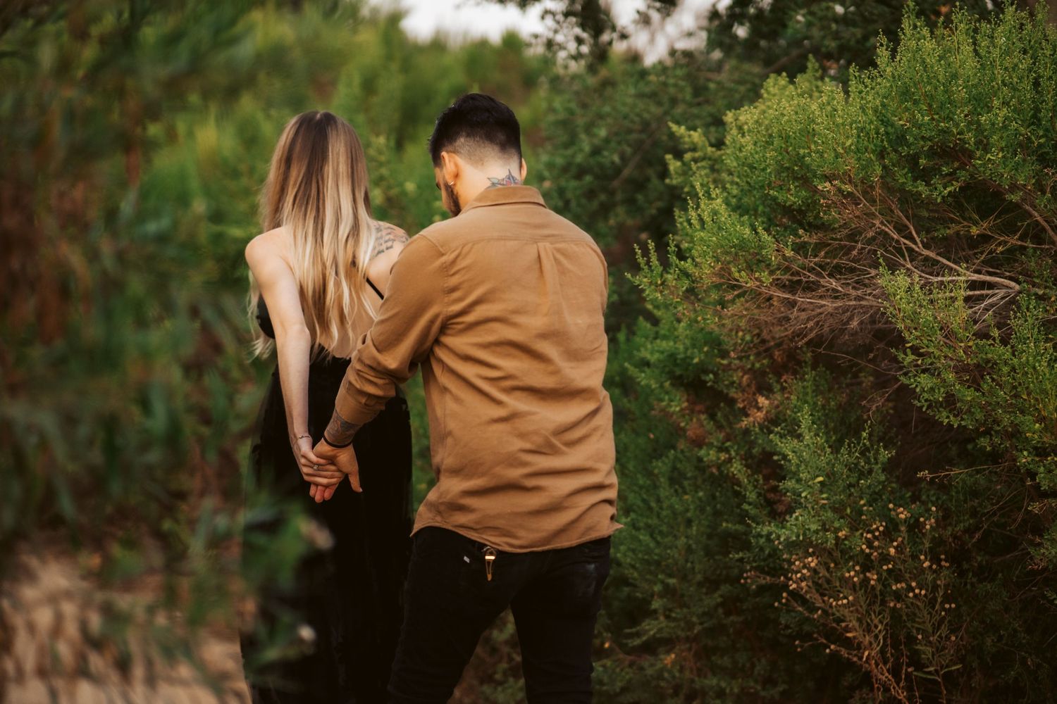 A couple in casual attire walks together through lush green foliage.
