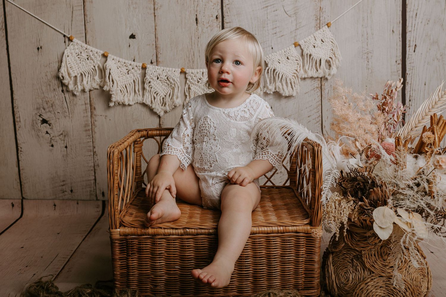 Baby in lace dress poses against distressed white wooden wall with dried floral accents.