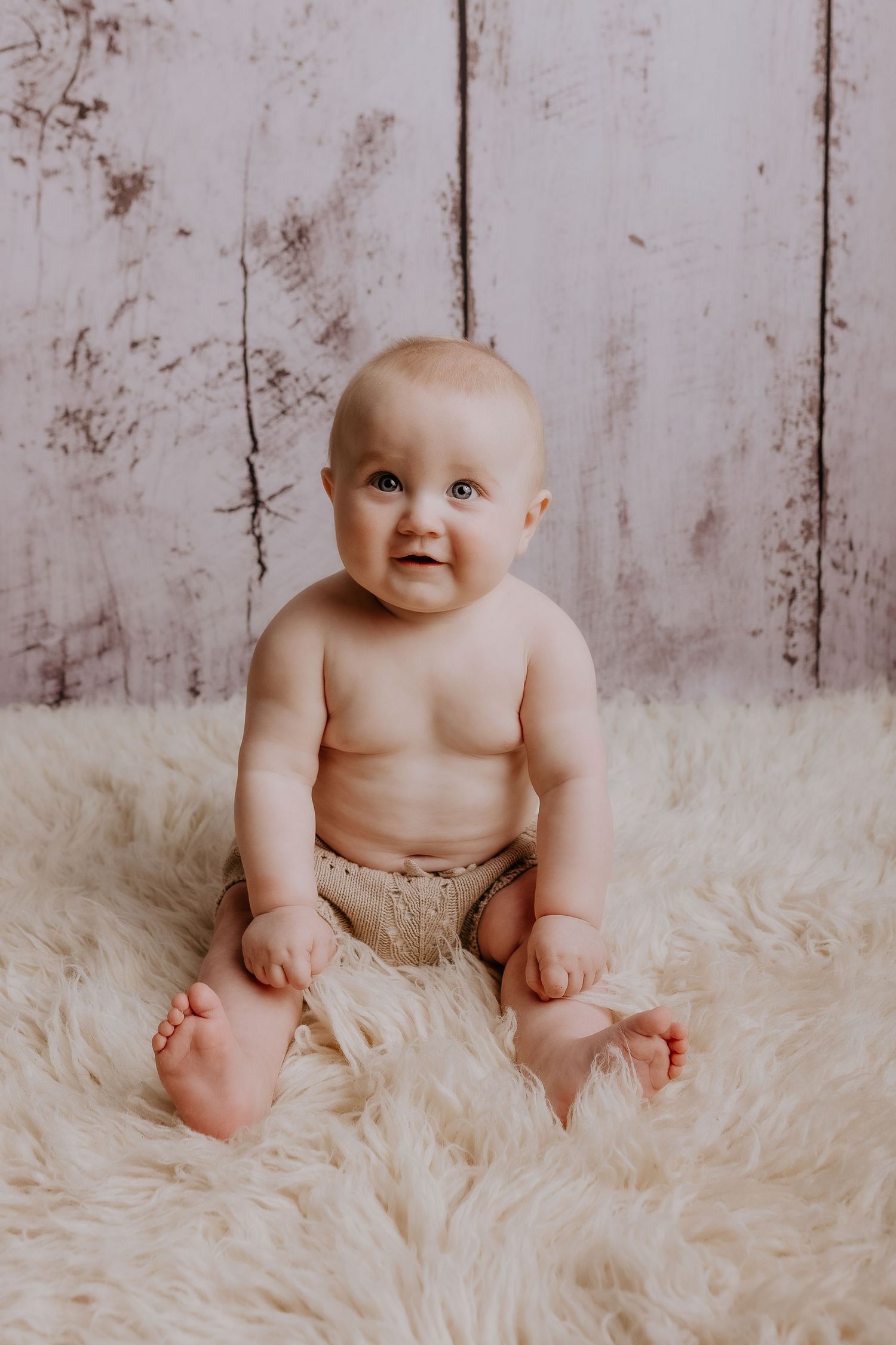 Baby sitting on fluffy white rug shows happy expression against distressed wood background.