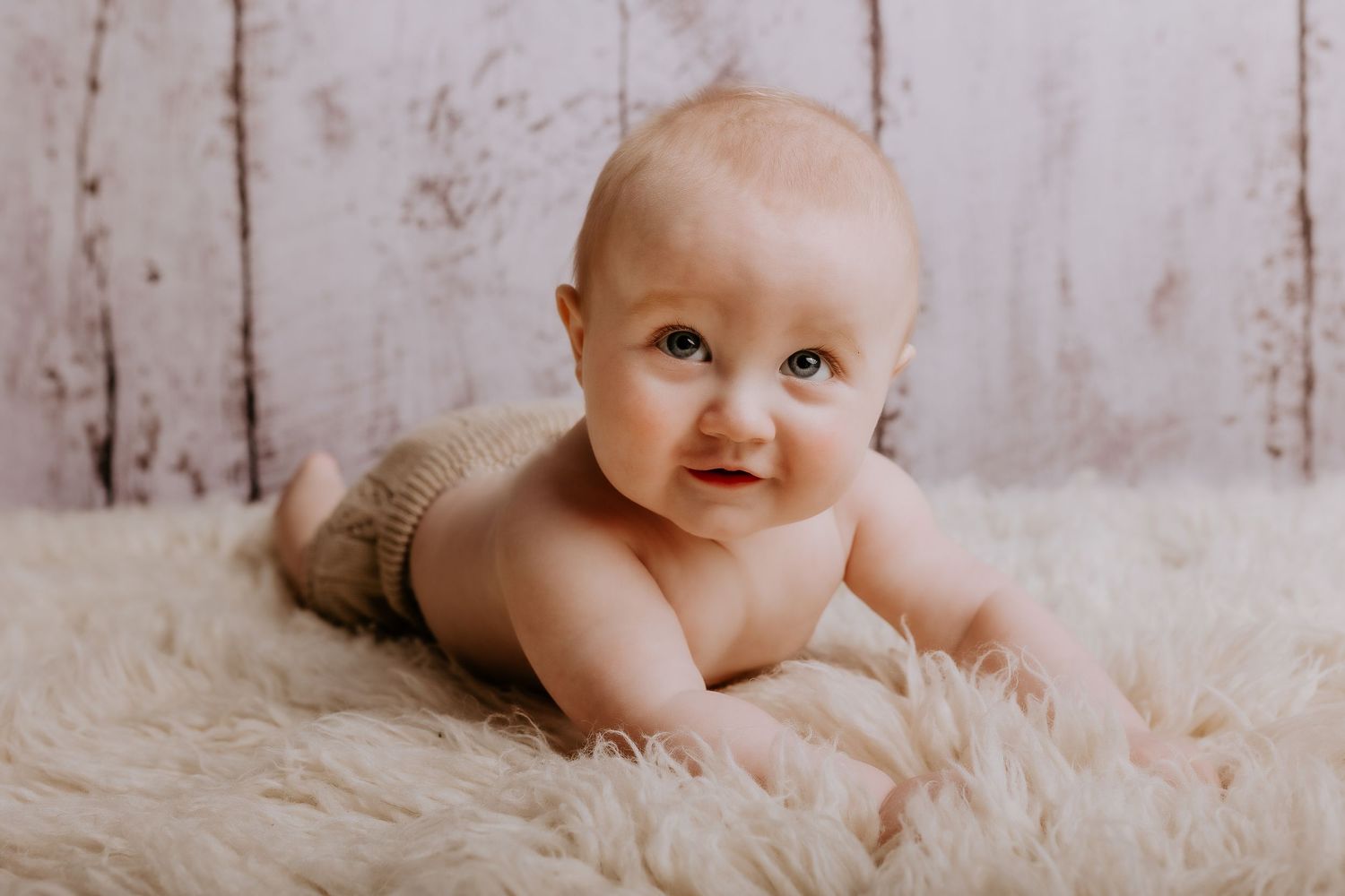 Baby poses on soft white fur blanket against rustic wooden wall background.