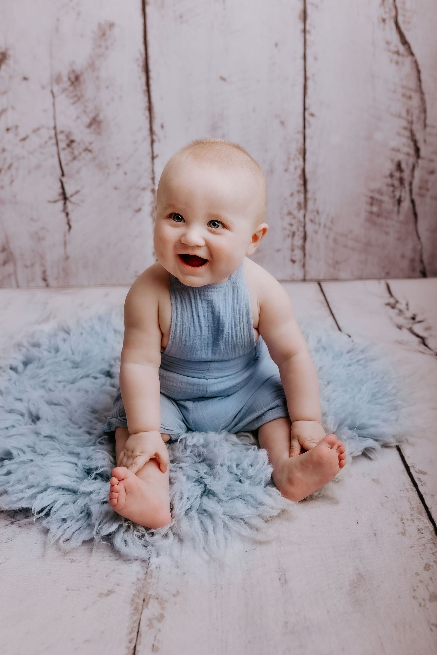 Baby in blue outfit smiling while sitting on soft blue rug during photo shoot.