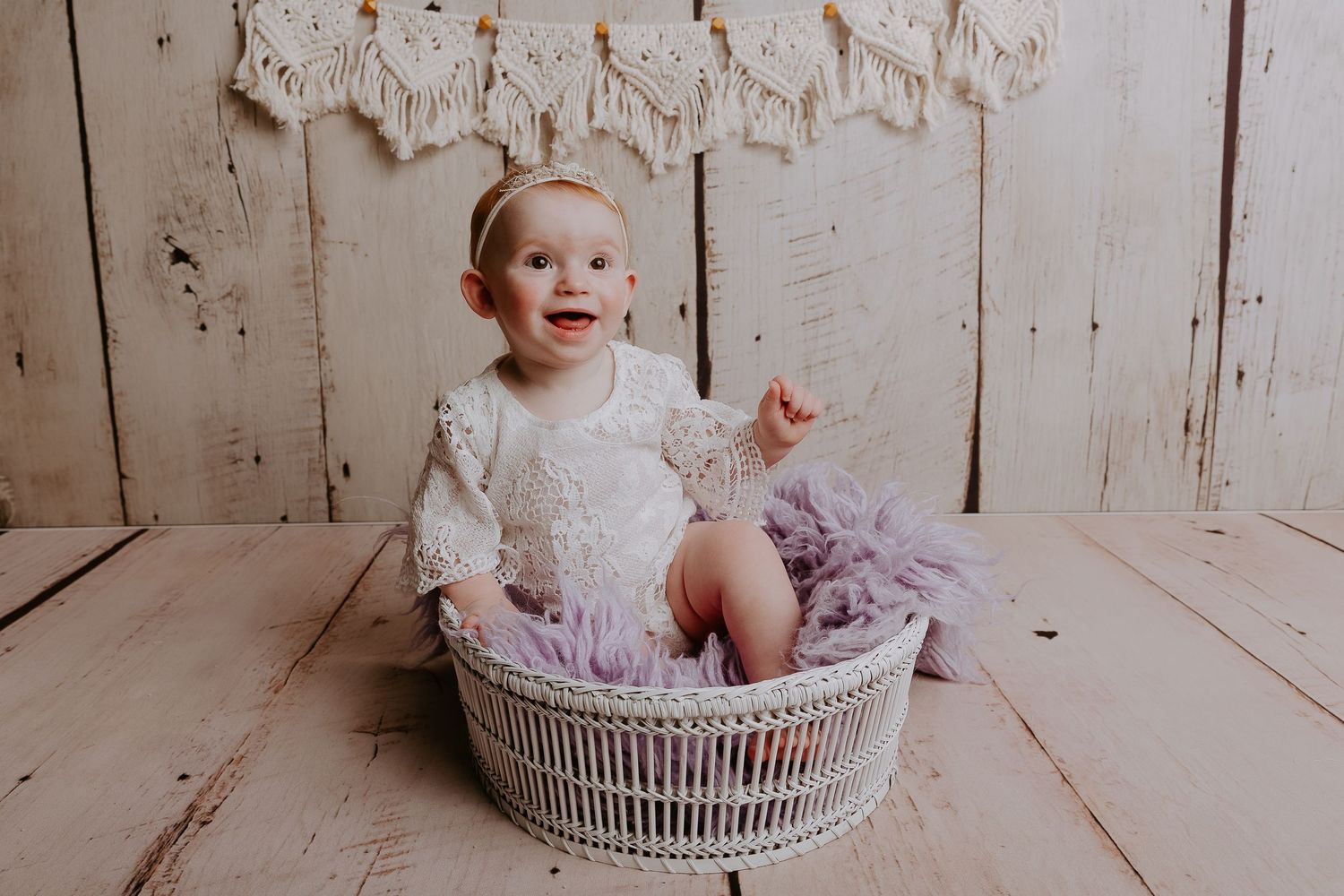 Baby poses in white lace dress inside decorative wicker basket against weathered wood wall.