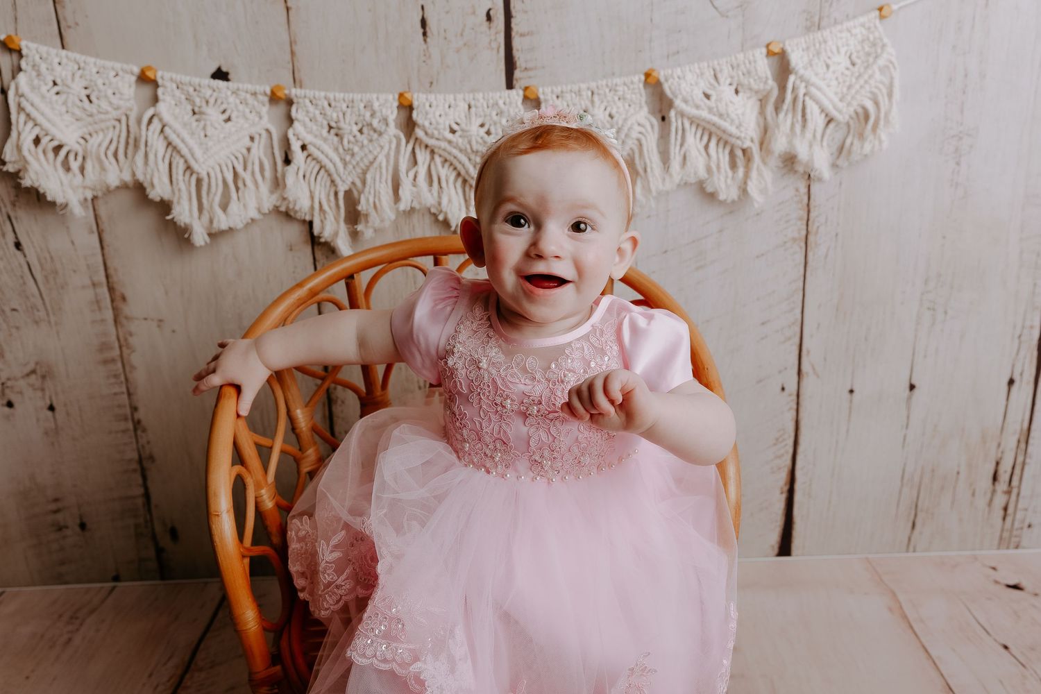 Baby wearing pink tulle dress sits in vintage wicker basket against rustic white backdrop.
