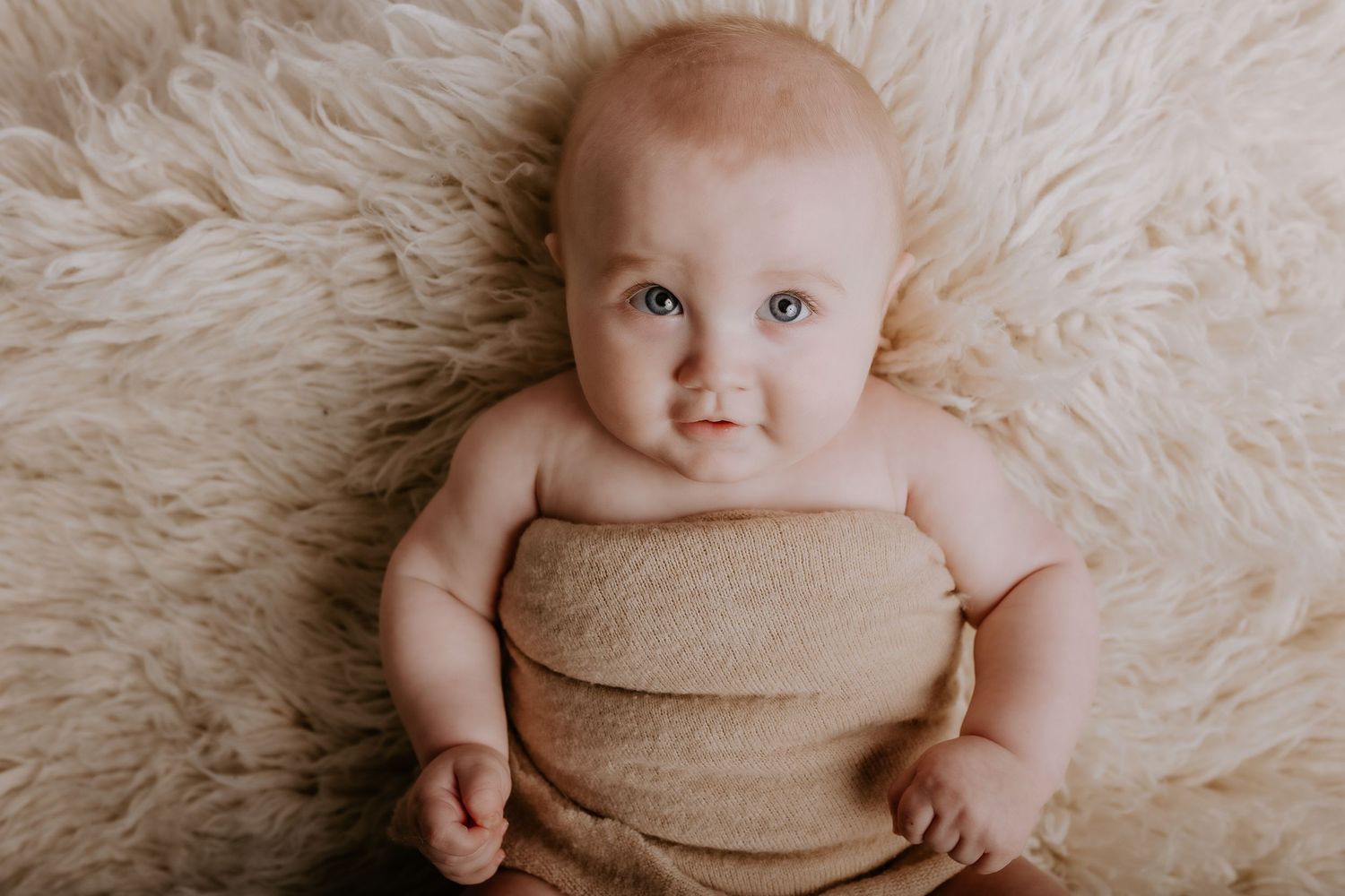 Baby wrapped in beige fabric lies on soft cream fur rug during professional photo shoot.