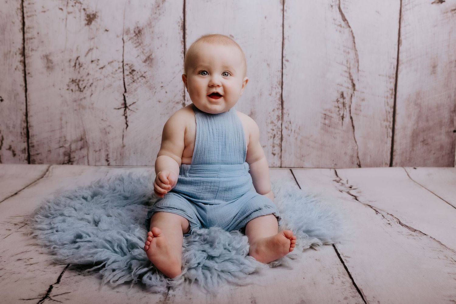 Baby wearing blue romper sits on fluffy rug during professional photography session.
