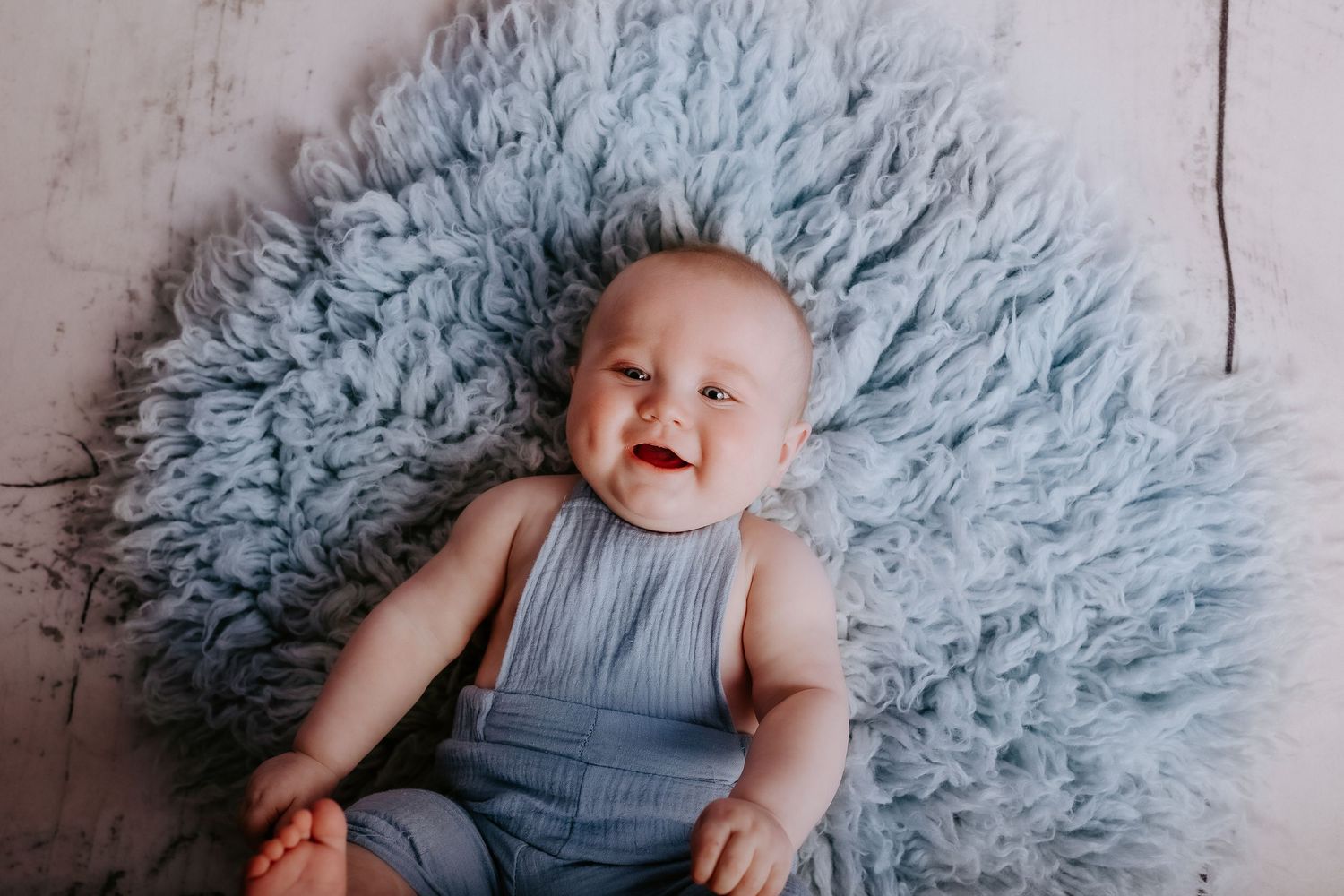 Baby wearing blue romper lying on fluffy blue rug against white wooden backdrop.