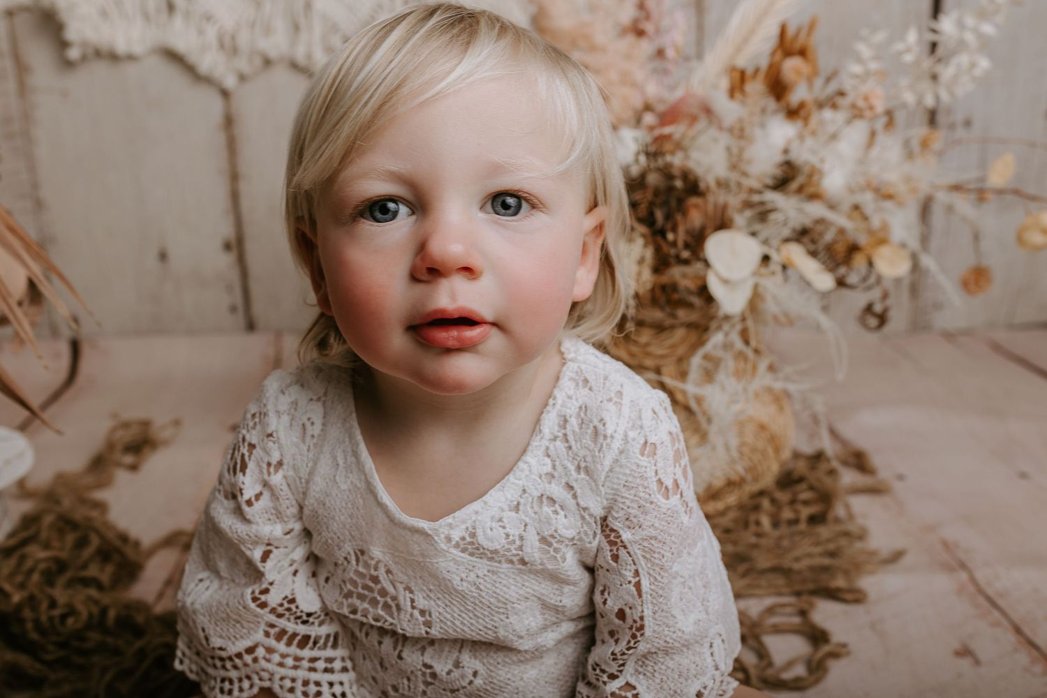 Baby in vintage lace outfit smiles during professional portrait session with rustic backdrop.