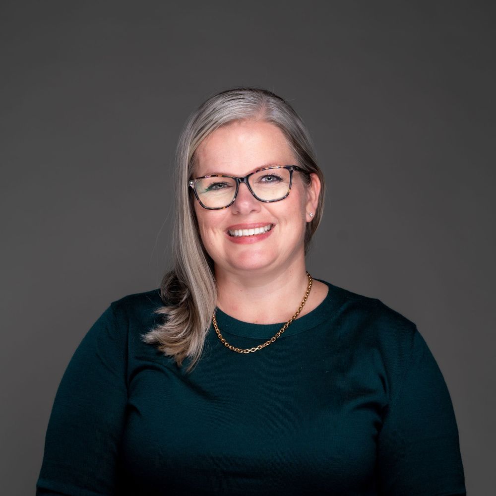 Professional headshot of person in dark teal sweater and glasses against gray background.