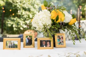 Elegant floral centerpiece with yellow roses and white hydrangeas next to framed photos.