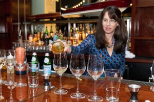 A bartender in a blue patterned top arranges wine glasses on a polished wooden bar counter.