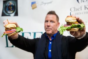 Close-up shot of man holding two gourmet burgers with both arms extended.