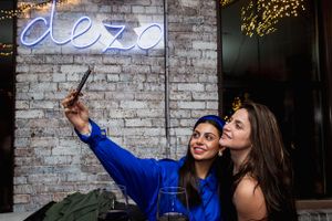 Two friends taking selfies in front of a neon sign at a trendy bar with exposed brick walls.