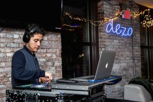 DJ performing with laptop and equipment under blue neon signage at night venue.
