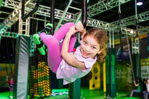 Young girl in pink outfit swinging on aerial equipment at indoor playground with green lighting.