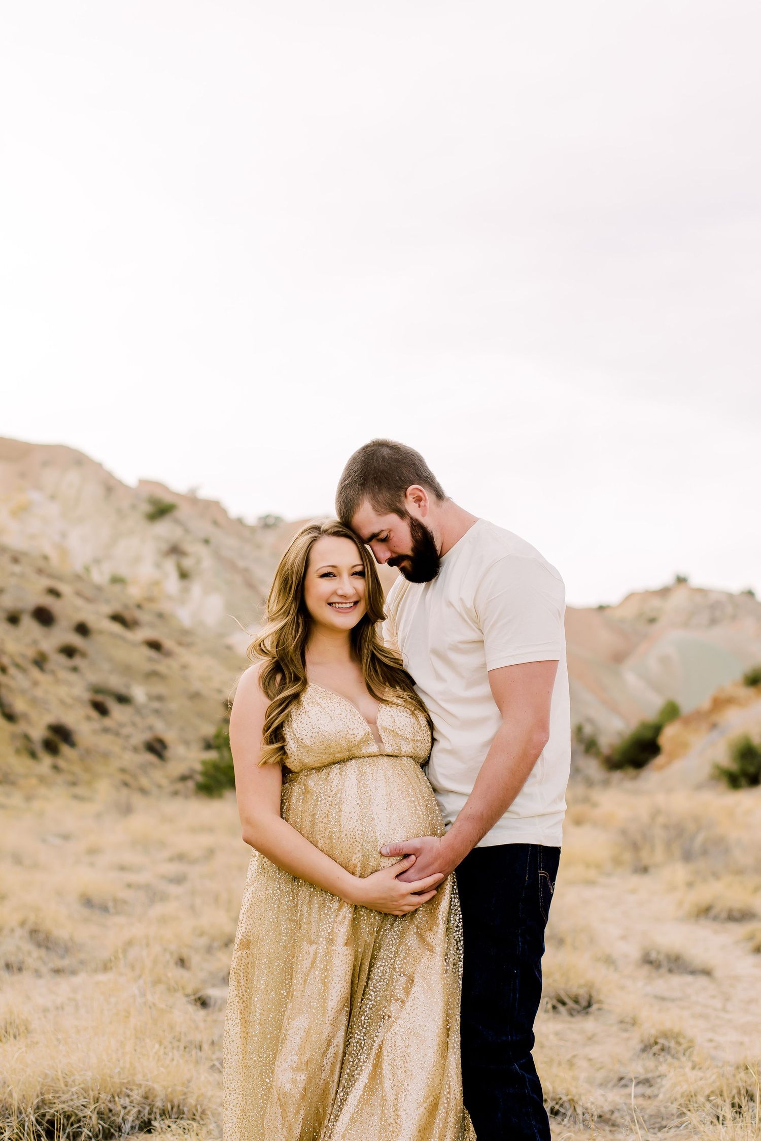 Expecting couple in desert landscape pictures with woman in gold dress and man in white shirt embracing against mountain backdrop.