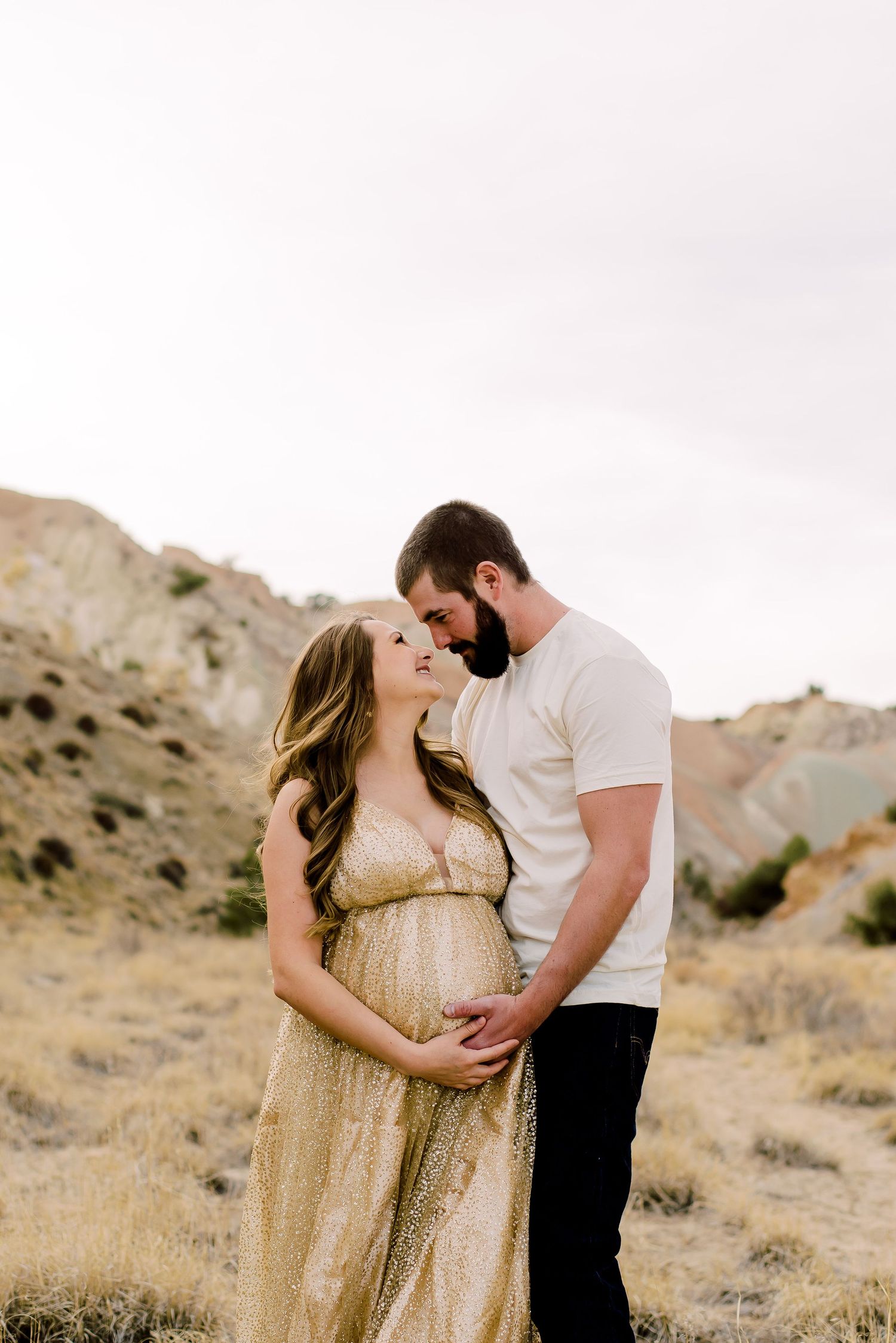 Pregnant couple embracing outdoors in desert landscape, woman in gold maternity gown, man in white tee and black jeans.