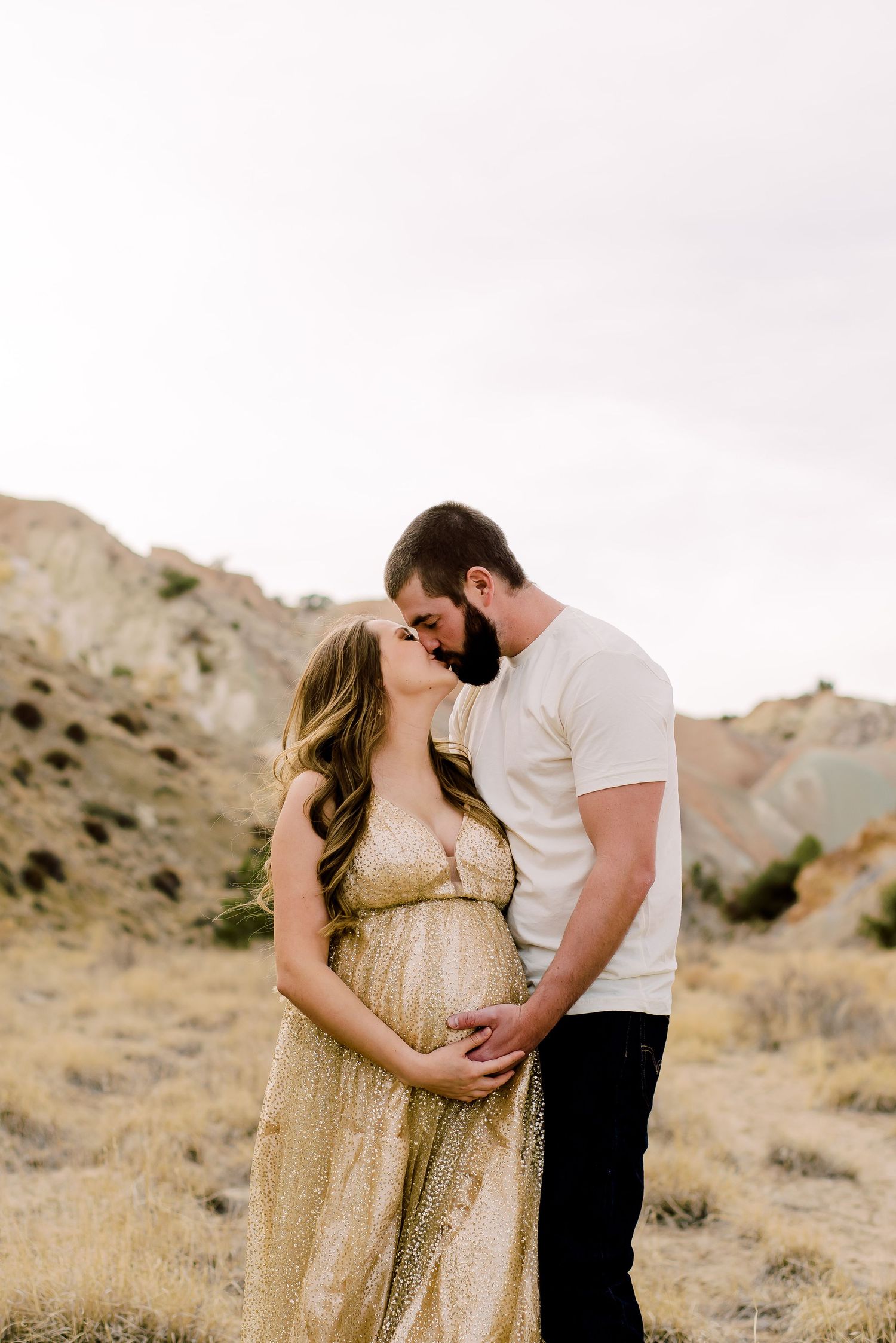 A couple stands in a romantic embrace on a desert hillside, with the woman wearing a flowing gold dress.