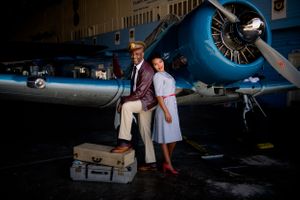 A vintage-styled couple poses with antique luggage in front of a classic airplane.