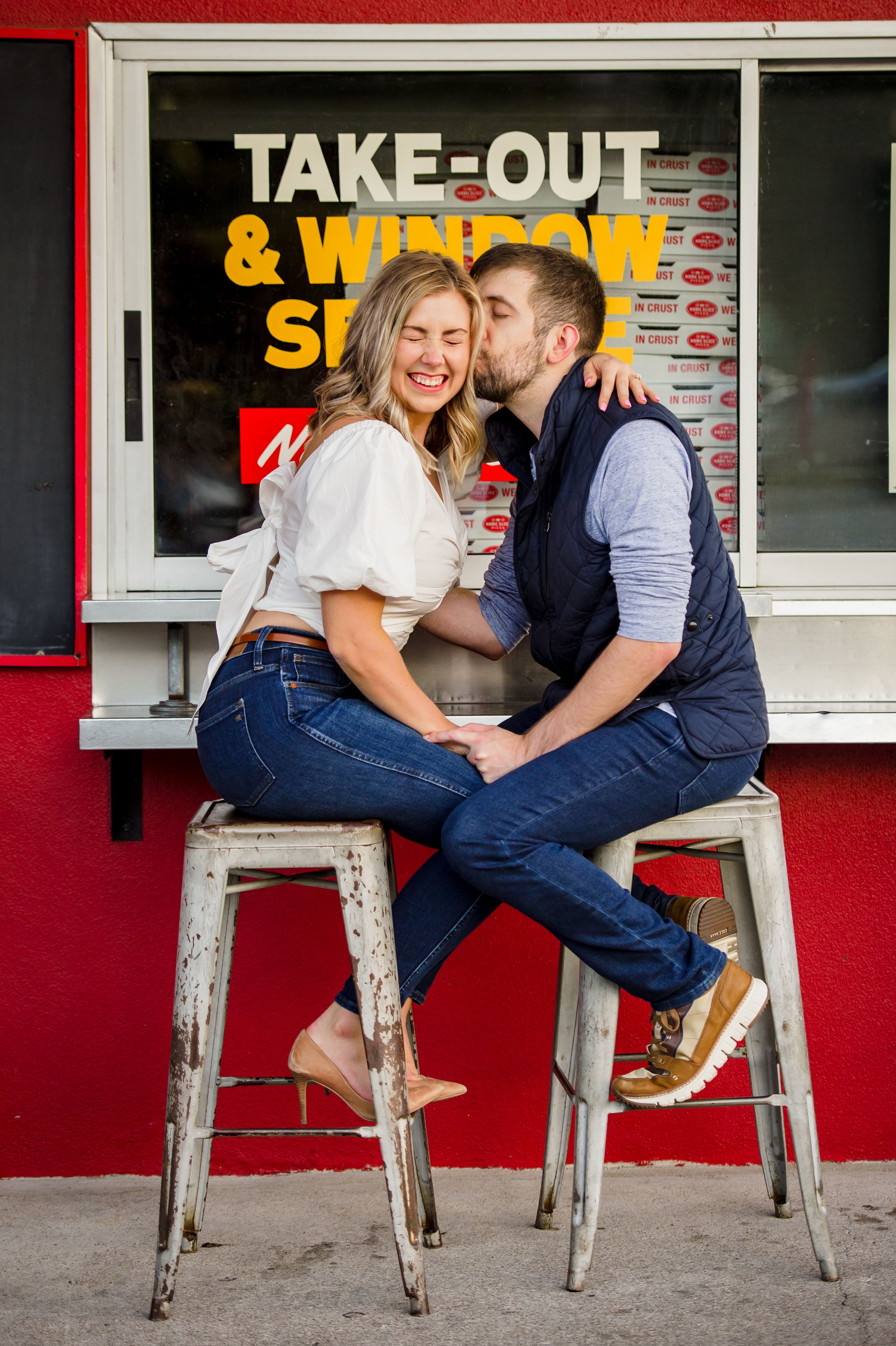 A couple shares a playful moment on metal stools outside a take-out restaurant window.