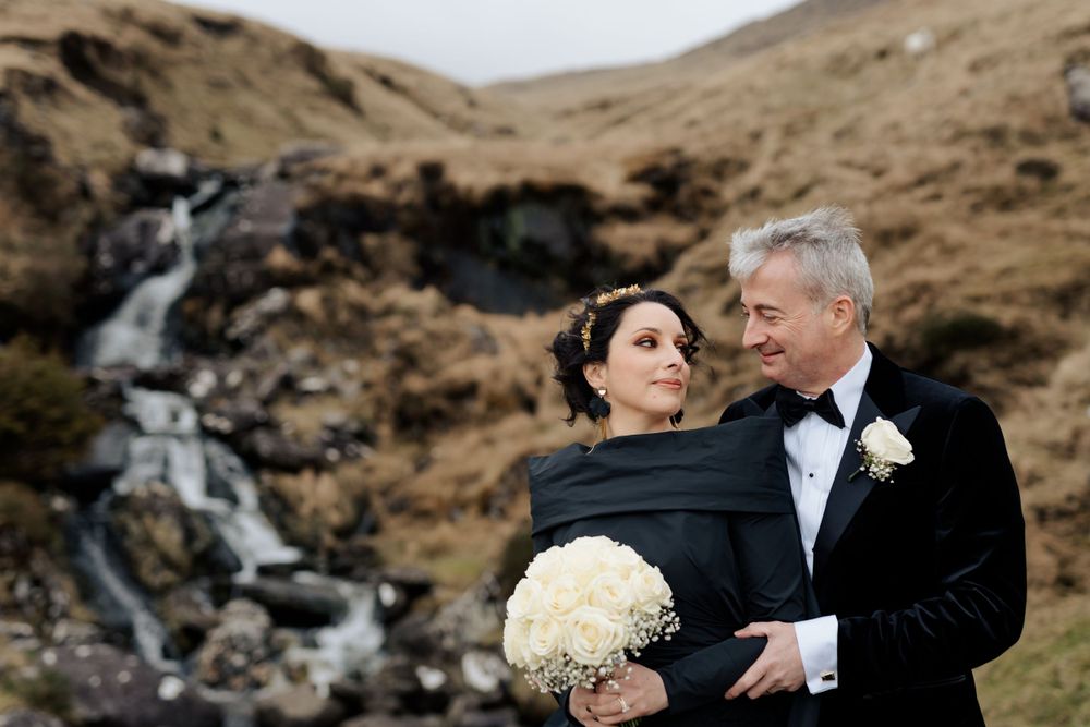 Newlyweds pose beside a scenic waterfall with a white bouquet in the Scottish highlands.