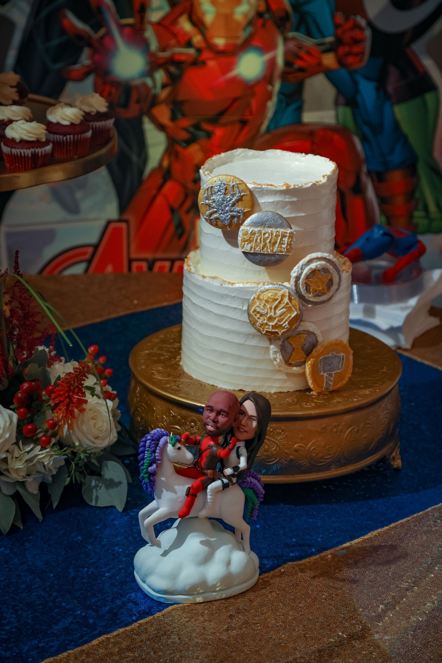 White wedding cake decorated with metallic cookie accents and unicorn figurine on blue tablecloth.