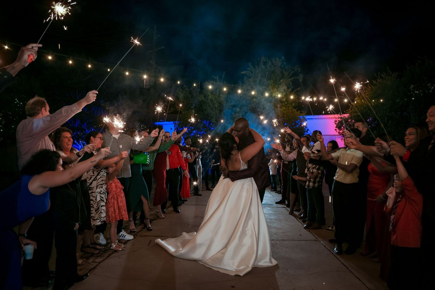 Newlyweds share a romantic dance under string lights while guests hold sparklers.