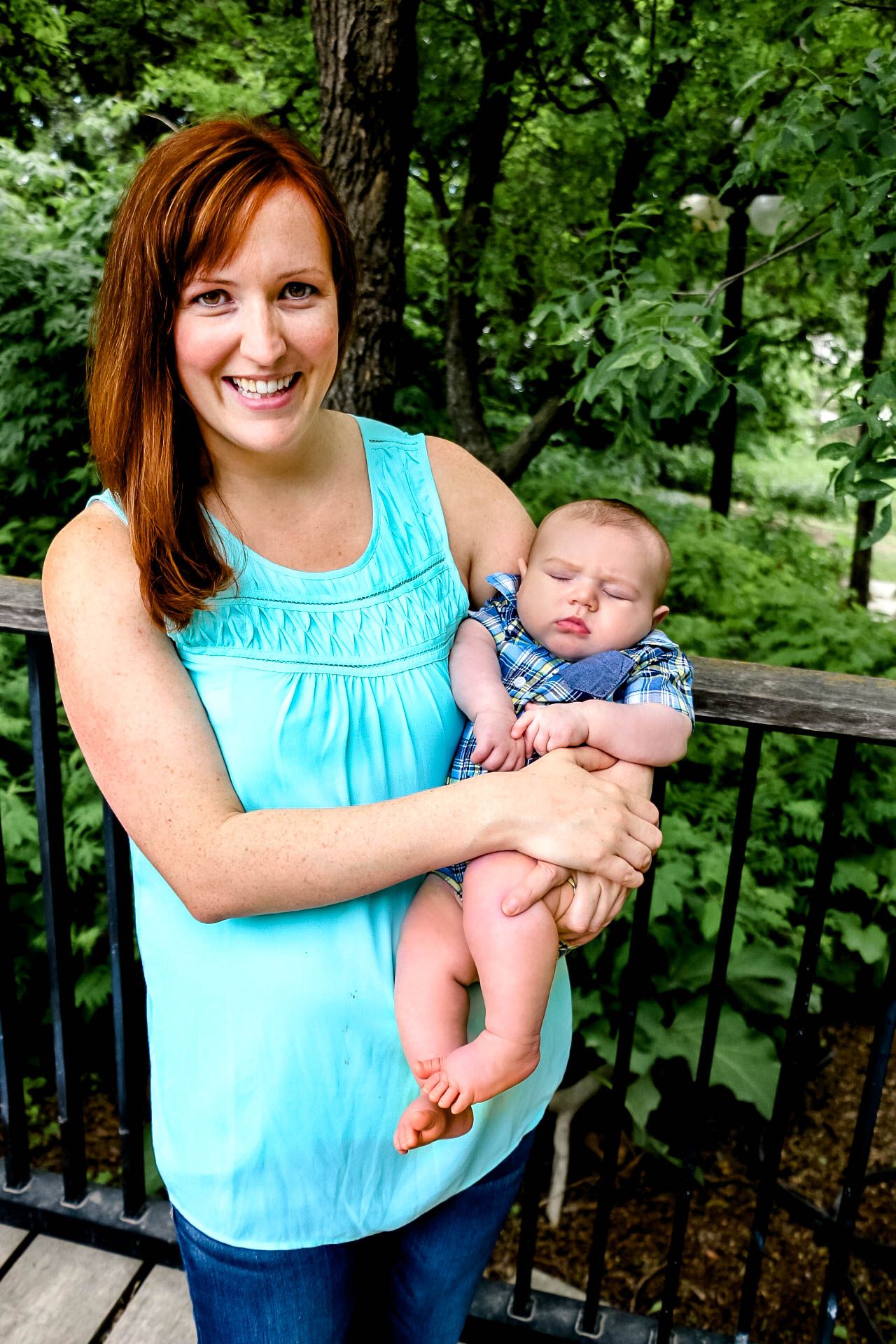 Woman in turquoise top holding sleeping infant on outdoor wooden deck surrounded by greenery.
