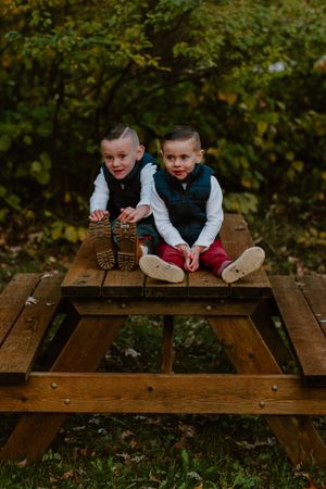Two children in matching vests sitting on a wooden picnic table in an autumn setting.